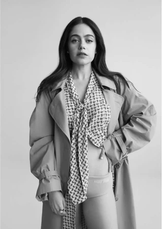 Monochrome portrait of a young woman wearing a trench coat, a patterned scarf, and branded underwear against a light background.
