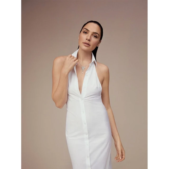 Portrait of Gal Gadot wearing a white button-down dress and necklace against a light beige background, with a shallow depth of field.