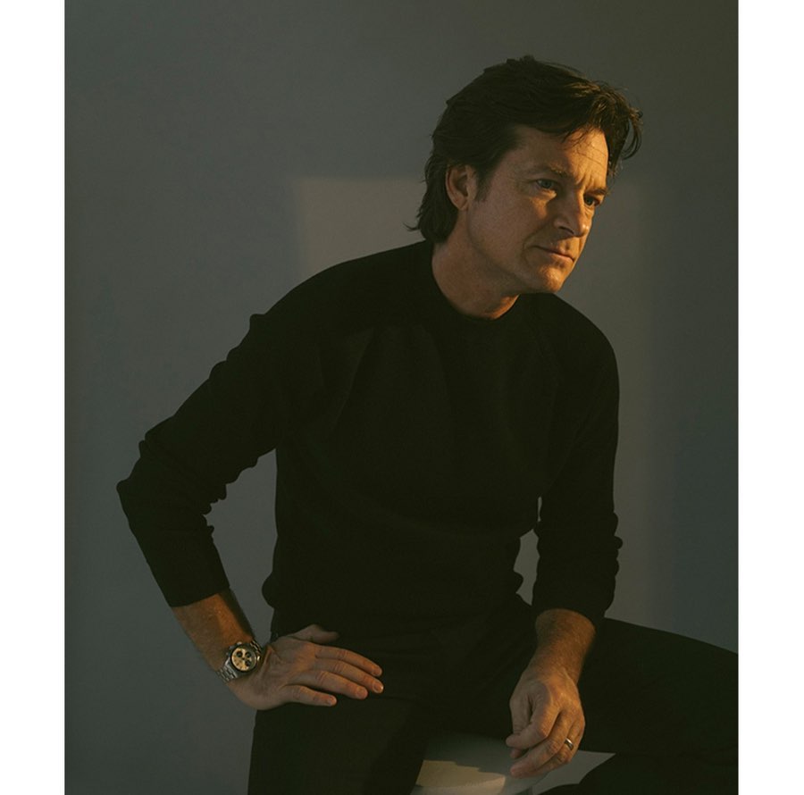 Portrait of Jason Bateman wearing a black sweater and sitting on a stool, looking off to the side in a photo studio.