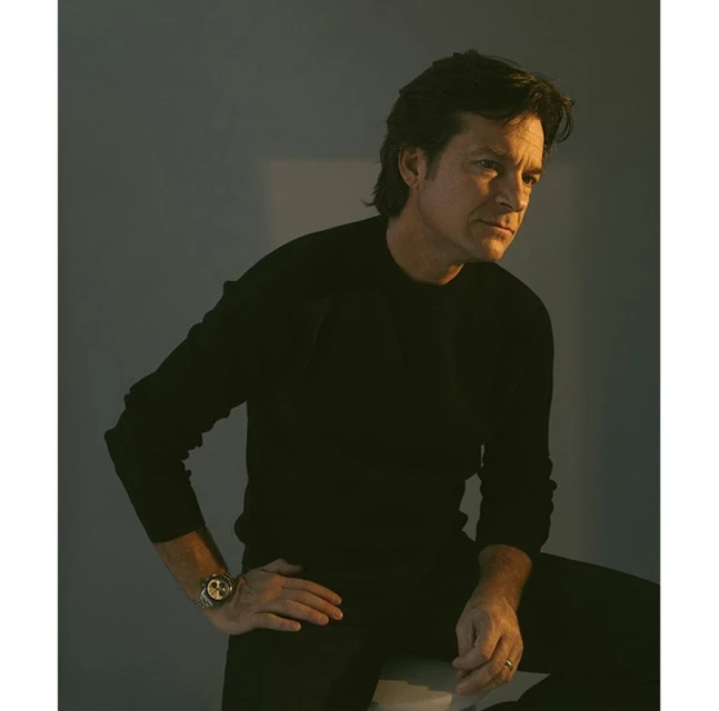 Portrait of Jason Bateman wearing a black sweater and sitting on a stool, looking off to the side in a photo studio.