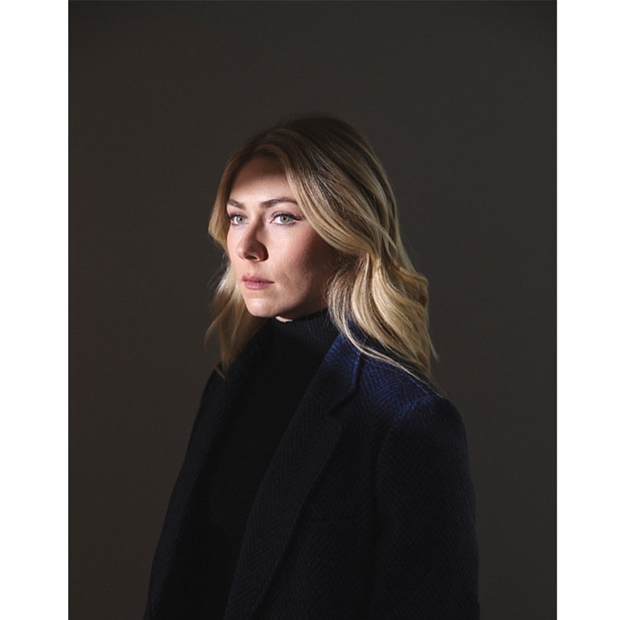 Portrait of a blonde woman in a black turtleneck and a navy coat against a dark grey background in a studio environment.