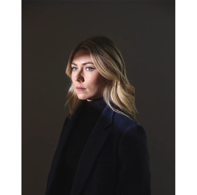 Portrait of a blonde woman in a black turtleneck and a navy coat against a dark grey background in a studio environment.