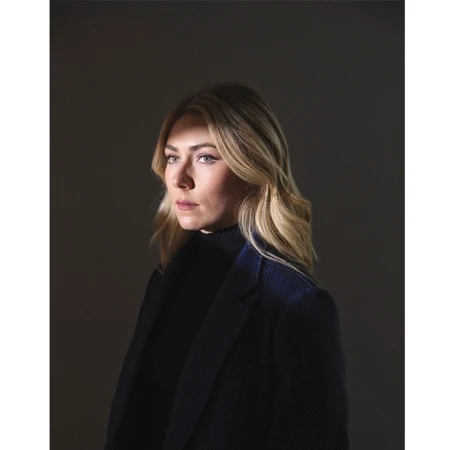 Portrait of a blonde woman in a black turtleneck and a navy coat against a dark grey background in a studio environment.