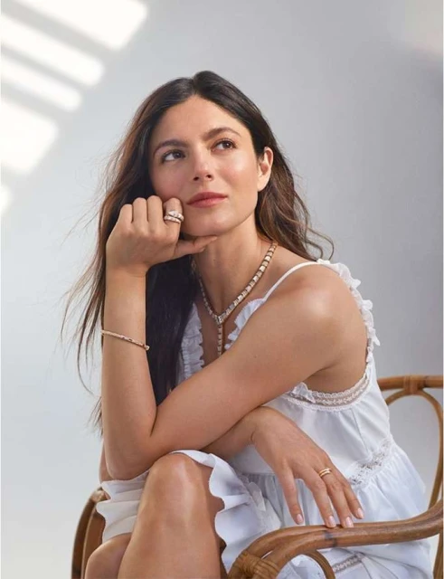 Woman wearing jewelry sits in a wicker chair, looking up with her hand near her face in a brightly lit indoor setting.