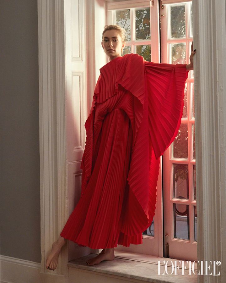 Woman in red pleated gown poses by window in fashion editorial, barefoot with neutral expression and hand on frame, magazine logo at bottom.