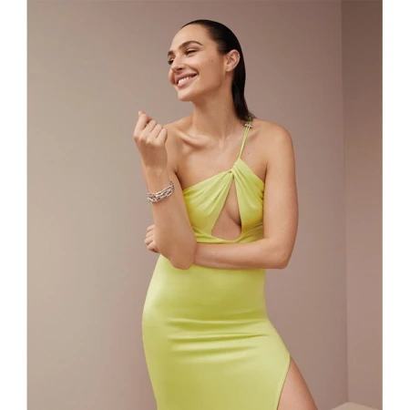 A woman with dark hair smiles while wearing a chartreuse dress, silver bracelet, and posing in a light, neutral-tone indoor setting.