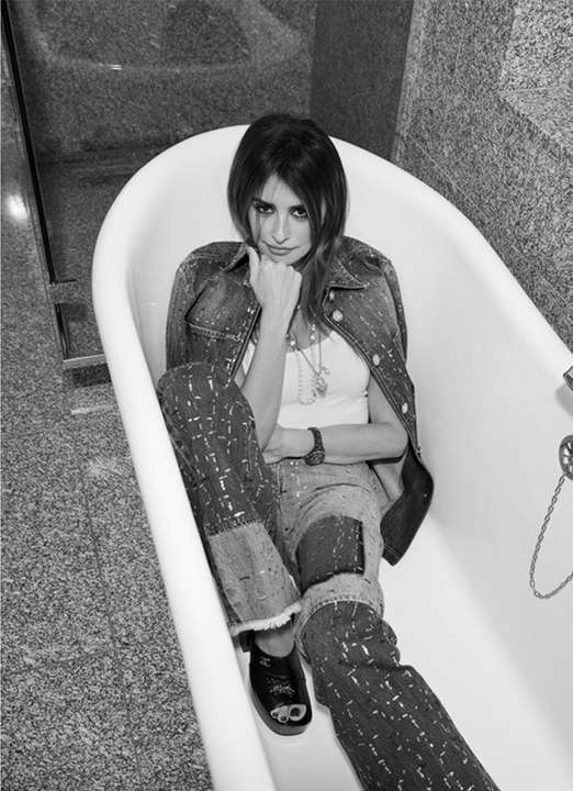 Black and white portrait of an adult woman in patterned jeans and denim jacket, sitting in a white bathtub with granite walls.