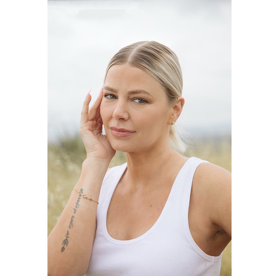 Blonde woman in white tank top poses outdoors with hand near face, set against blurred field and cloudy sky backdrop in portrait format.