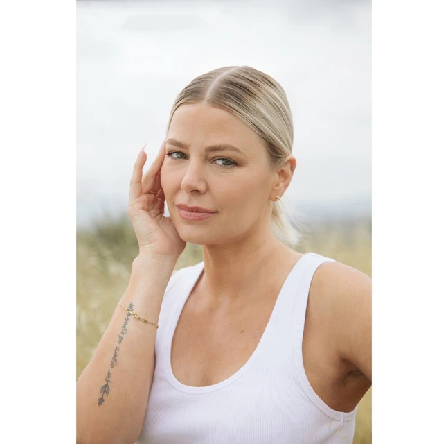 Blonde woman in white tank top poses outdoors with hand near face, set against blurred field and cloudy sky backdrop in portrait format.
