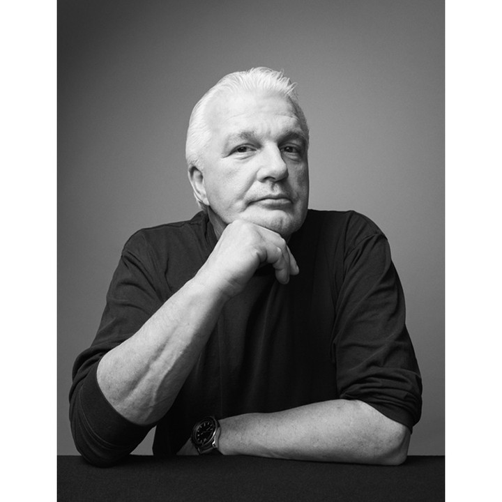 Portrait of an elderly white man resting chin on hand wearing a dark long-sleeve shirt against a grey seamless background