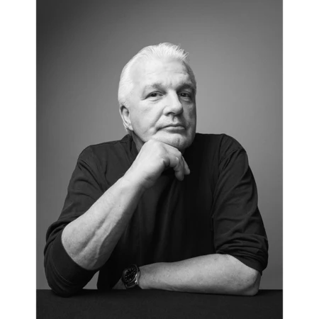Portrait of an elderly white man resting chin on hand wearing a dark long-sleeve shirt against a grey seamless background