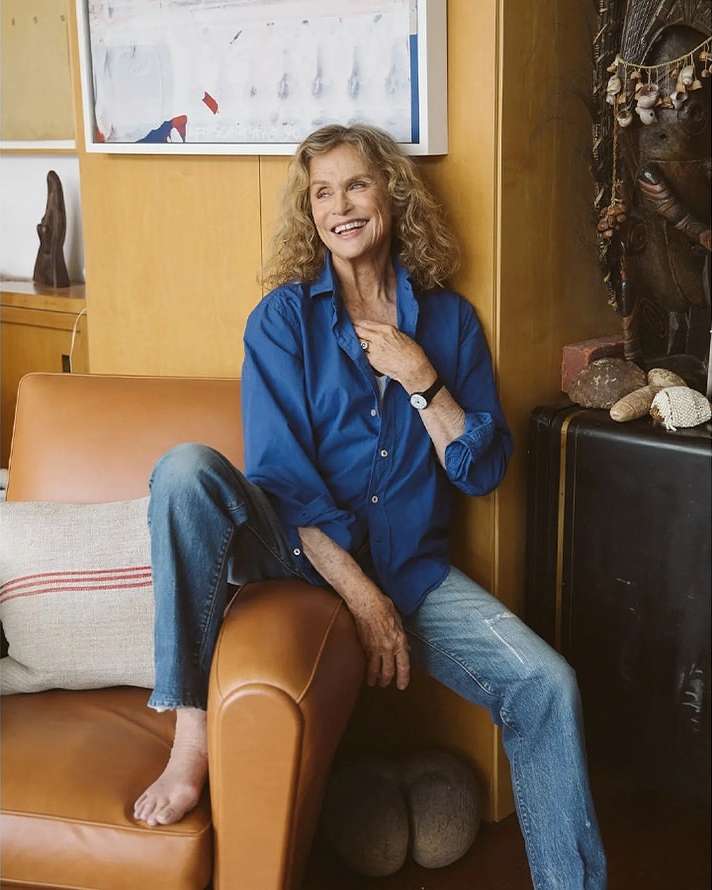 Older woman with curly hair sits smiling on a tan chair wearing a blue button-down shirt and jeans, with an art piece in the background.