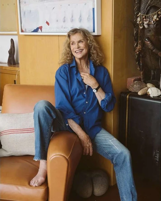 Older woman with curly hair sits smiling on a tan chair wearing a blue button-down shirt and jeans, with an art piece in the background.