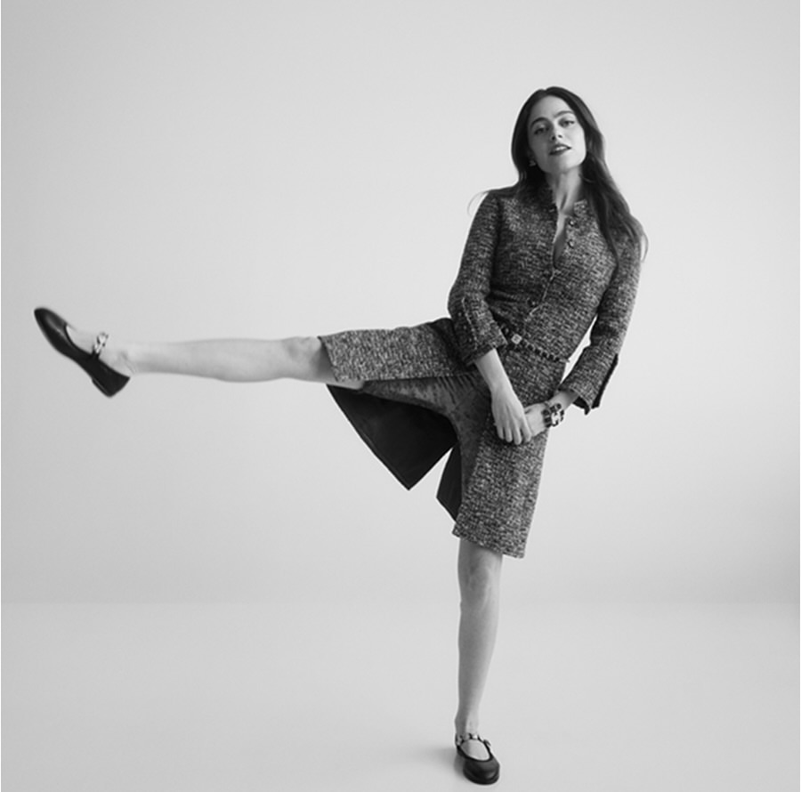 A black and white fashion shot of a woman in a textured suit and black flats striking a playful pose against a plain background.