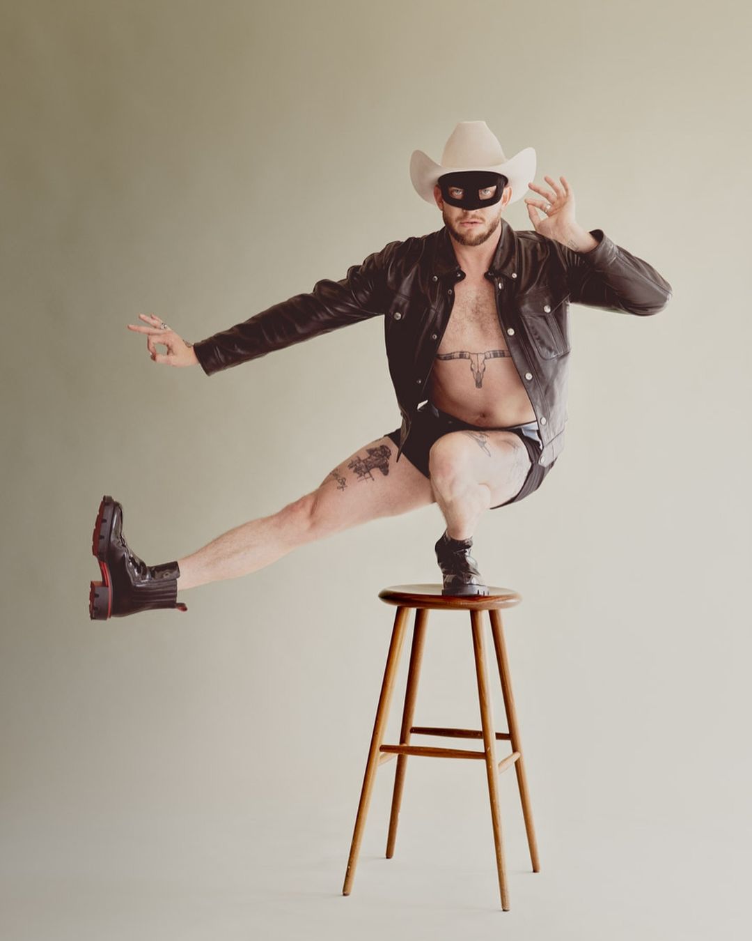 Man wearing a cowboy hat, eye mask, and leather jacket balancing on one leg on a wooden stool.
