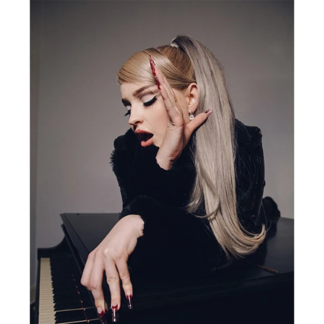 Blonde woman with ponytail, heavy eye makeup, and long red nails poses with her hands near a black piano in a studio setting.