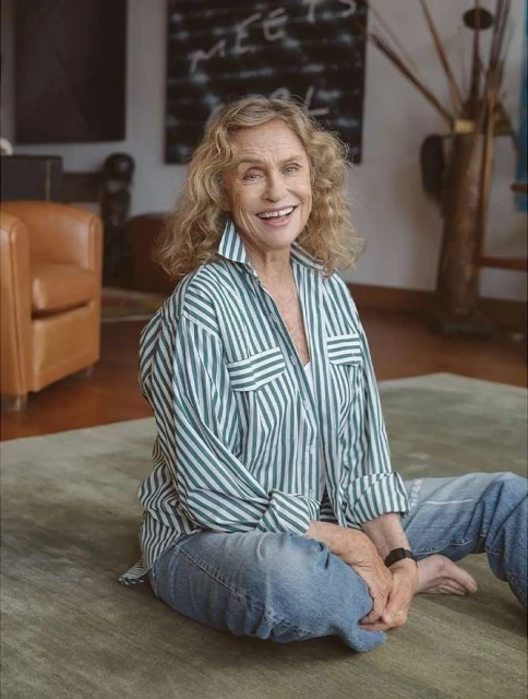Smiling mature woman sits on the floor in an open striped shirt and jeans in a living room setting. Casual and comfortable style.