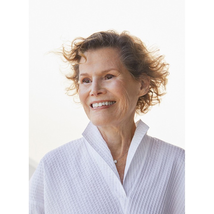Headshot of a smiling older woman with short curly hair, wearing a white bathrobe and a pearl necklace, set against a bright background.