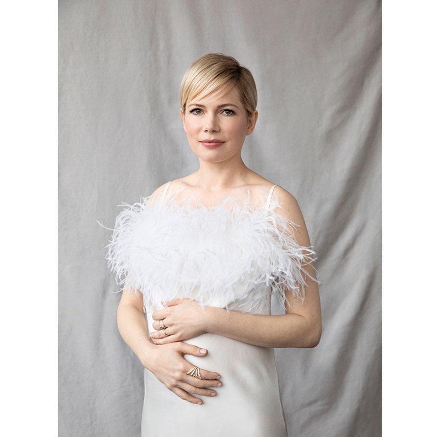 Michelle Williams poses in a white gown with feathered detail against a grey backdrop. Portrait of the actress.