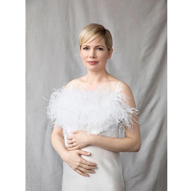 Michelle Williams poses in a white gown with feathered detail against a grey backdrop. Portrait of the actress.