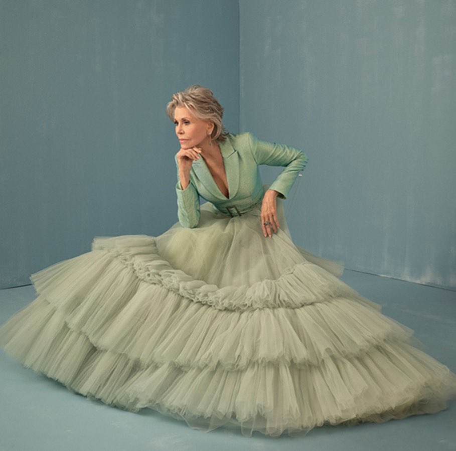 Jane Fonda seated wearing a sage green blazer and voluminous tiered tulle skirt against a light blue studio backdrop.