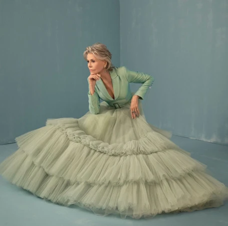Jane Fonda seated wearing a sage green blazer and voluminous tiered tulle skirt against a light blue studio backdrop.
