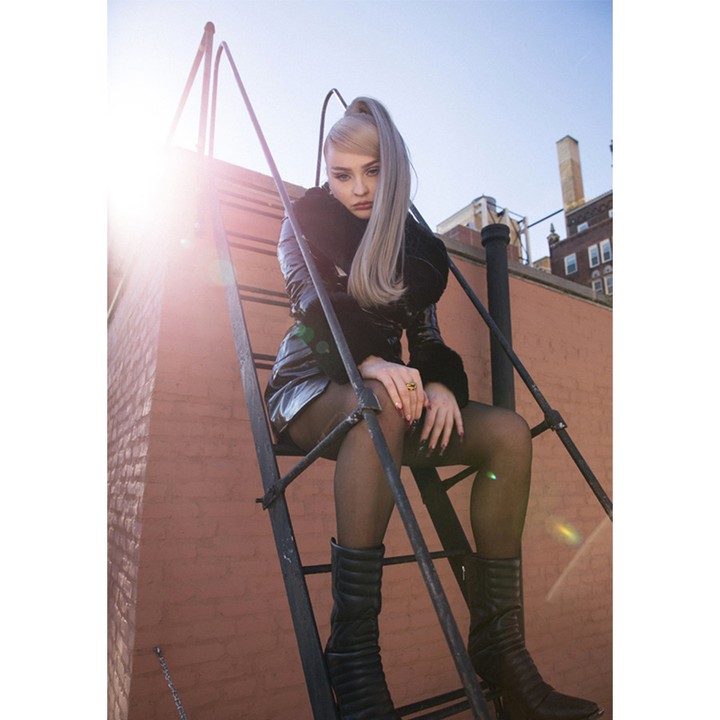 Young woman with ponytail wearing a jacket and skirt sits on a metal staircase outdoors with an orange brick building background.