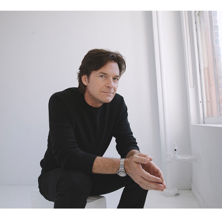 Adult male poses sitting on a white stool in a minimalist studio. He wears a black top and pants with a silver wristwatch.