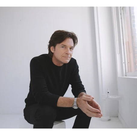 Adult male poses sitting on a white stool in a minimalist studio. He wears a black top and pants with a silver wristwatch.