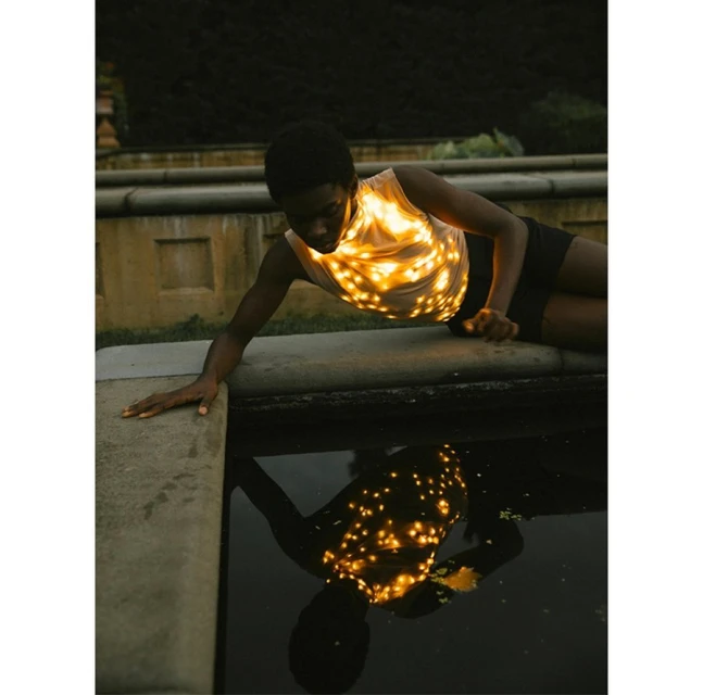 Woman in tank top with light patterns lying on stone ledge with water reflection in outdoor setting at dusk. Calm pose. Fashion.