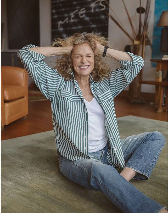Smiling mature woman with curly blonde hair sitting cross-legged on a rug in a casual striped shirt and jeans in a brightly lit room.