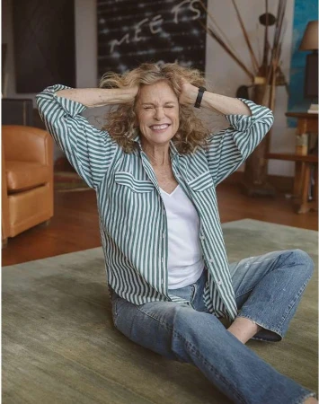Smiling mature woman with curly blonde hair sitting cross-legged on a rug in a casual striped shirt and jeans in a brightly lit room.