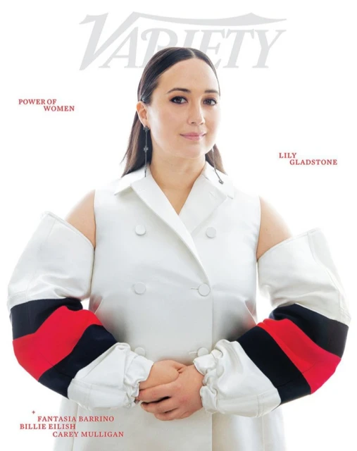 Lily Gladstone graces the cover of Variety magazine, wearing a white coat with red and black sleeve accents, in a studio setting.