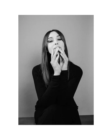 Portrait of Monica Bellucci wearing a black turtleneck with hands near her face against a grey backdrop