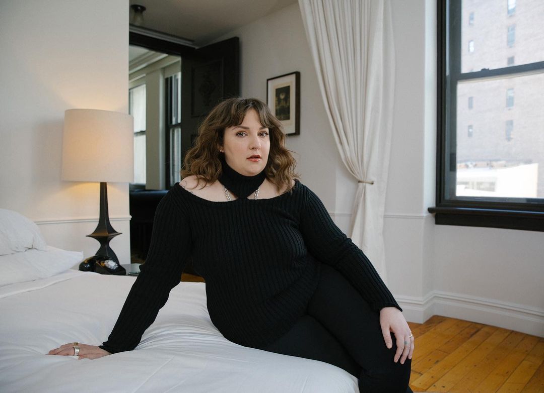 Portrait of a woman in a black top and pants sitting on a white bed in a naturally lit room, posing for a fashion shoot.