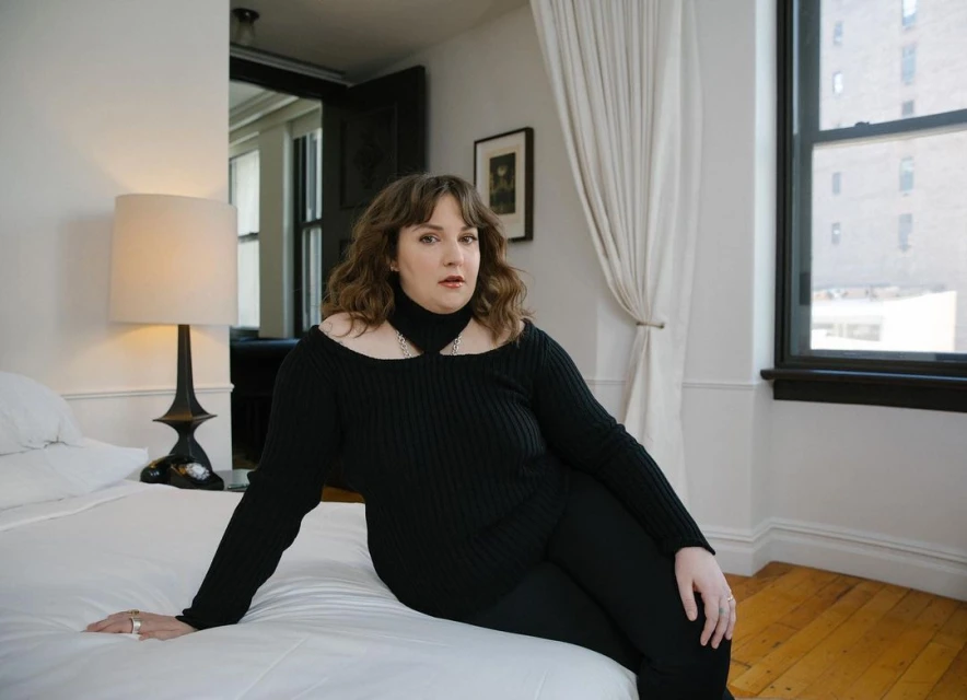 Portrait of a woman in a black top and pants sitting on a white bed in a naturally lit room, posing for a fashion shoot.