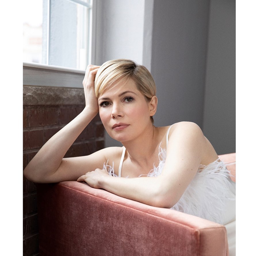 Portrait of Michelle Williams with short blonde hair, wearing a white top, seated in a pink chair, next to a window.