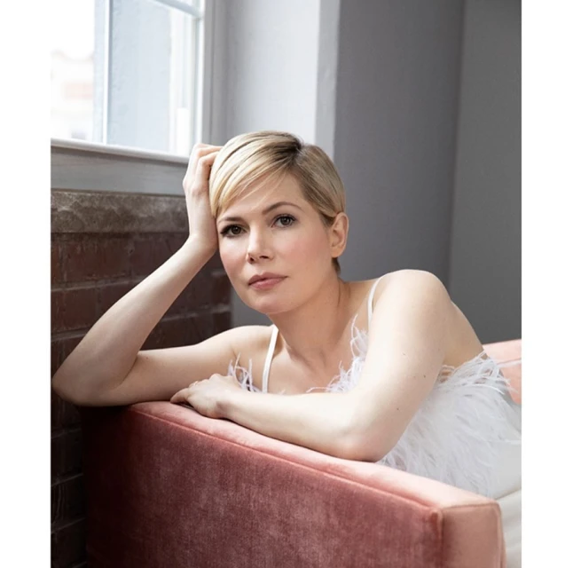 Portrait of Michelle Williams with short blonde hair, wearing a white top, seated in a pink chair, next to a window.