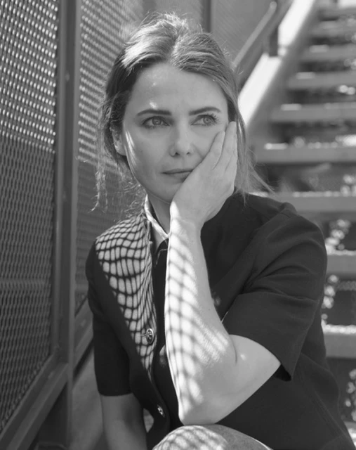 Black and white portrait of a woman with hand on face sitting on metal steps, wearing a button-down shirt and patterned top layer.
