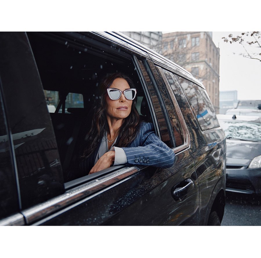 Adult woman in a pinstripe blazer and sunglasses in a black car looking out the window in an urban environment with buildings.