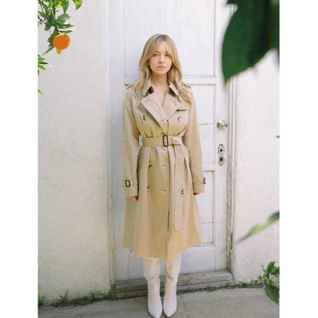 Blonde woman in trench coat and white boots poses near white door with orange tree in background. Fashion portrait in daytime.