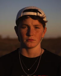 Portrait of a young man wearing a white cap and black t shirt with layered chains under golden hour light by Celeste Sloman