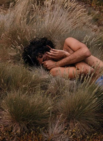 A person with dark curly hair lies curled up among tall dry grass in a field, touching their face.