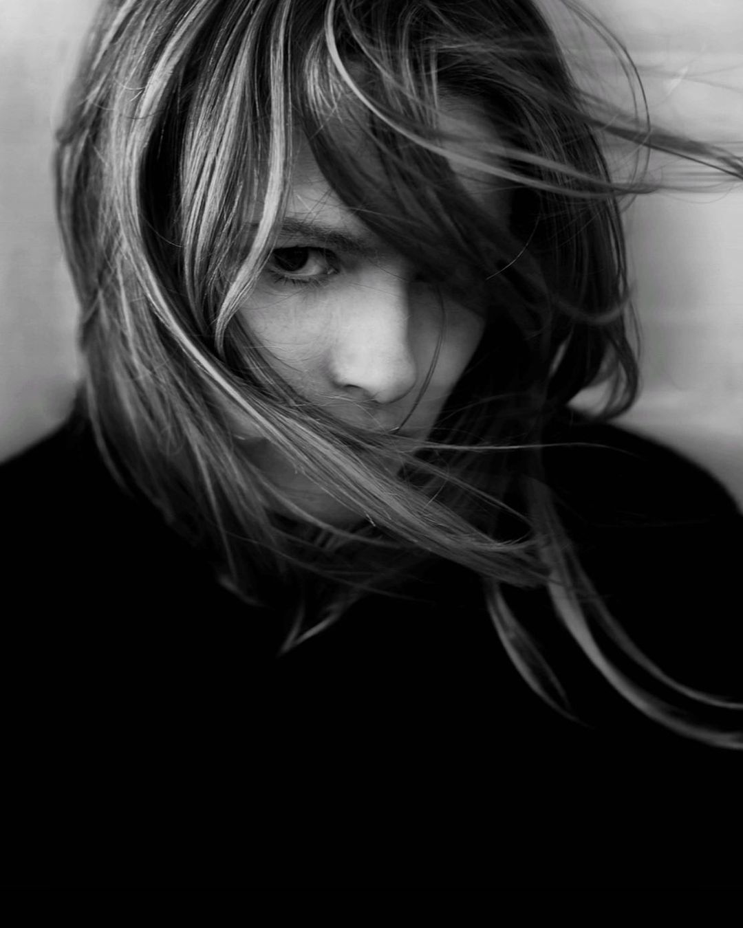 Black and white portrait of a person with shoulder-length hair covering part of their face, looking directly at the camera.