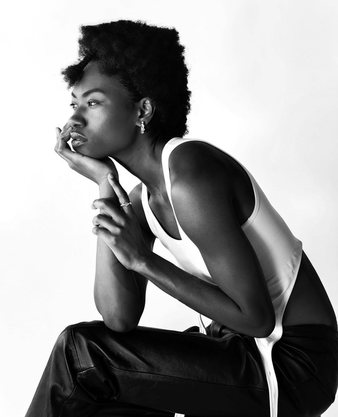 Monochromatic portrait of a Black model in a white tank top and black pants, sitting with hand to face, looking off to the side.