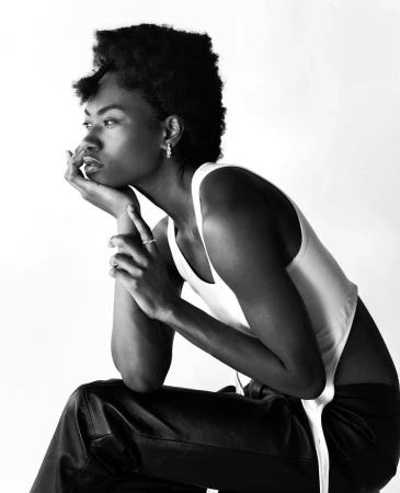Monochromatic portrait of a Black model in a white tank top and black pants, sitting with hand to face, looking off to the side.