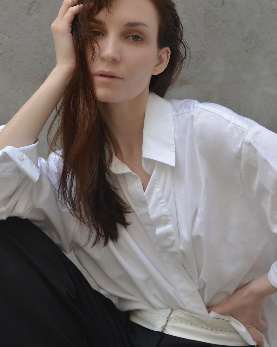 Portrait of a woman with long dark hair wearing a white collared shirt and black trousers against a neutral wall