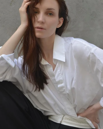 Portrait of a woman with long dark hair wearing a white collared shirt and black trousers against a neutral wall