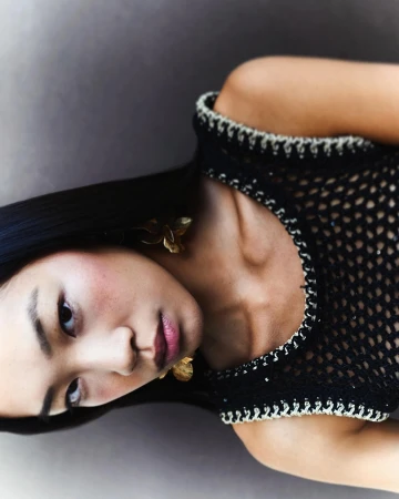 Close-up portrait of a woman wearing a black crocheted sleeveless top and gold earrings against a neutral grey background