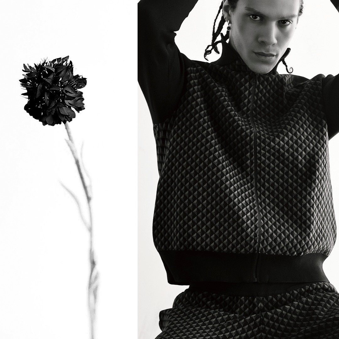 Studio shot of a black flower in soft focus next to a male fashion model in black, geometric-patterned athletic wear on a bright set.